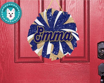Peut inclure: Un nœud de pom-pom de cheerleader bleu, blanc et or avec le nom "Emma" en lettres dorées. Le nœud est accroché à une porte rouge.