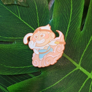 May include: A peach-colored enamel pin featuring a cartoon monkey with a staff, wearing a blue and white outfit, and sitting on a cloud. The monkey has a mischievous expression and is holding a staff.