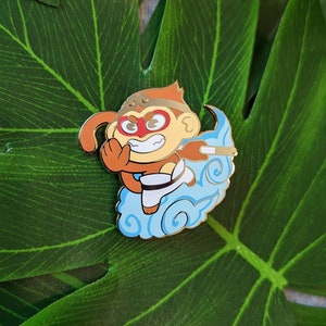 May include: A gold-colored enamel pin featuring a cartoon monkey wearing a red mask and holding a staff. The monkey is flying on a blue cloud with white swirls.