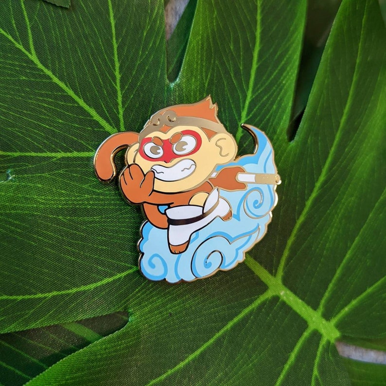 May include: A gold-colored enamel pin featuring a cartoon monkey wearing a red mask and holding a staff. The monkey is flying on a blue cloud with white swirls.