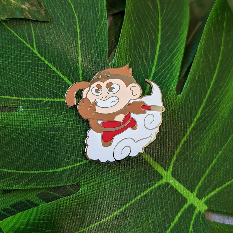 May include: Gold-colored enamel pin featuring a cartoon monkey in a red and white outfit, flying on a white cloud with a red and white staff. The monkey has a brown face with a mischievous expression.