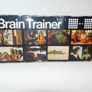 May include: Vintage "Brain Trainer" game box with a black background and white text. The box features multiple small images depicting various scenes. The game box is a retro item, likely from the mid-20th century.