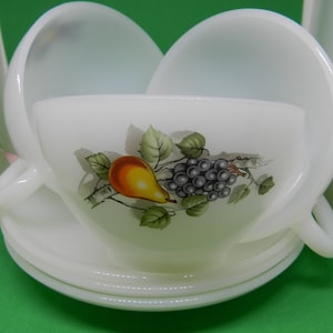 Op de afbeelding: Witte kopjes en schotels met een fruitontwerp. De kopjes hebben een oor en zijn versierd met een peer, druiven en groene bladeren. De schotels zijn rond en stapelbaar.