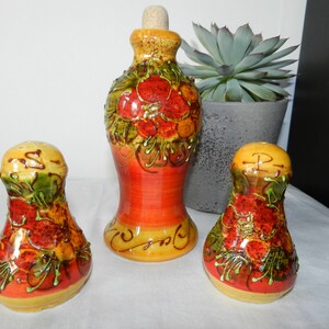Op de afbeelding: Een keramische set met een fles en twee zout- en peperstrooiers. De set heeft een levendig ontwerp van rode, oranje en groene bloemmotieven op een gele basis. De fles heeft een kurk. De set is waarschijnlijk voor gebruik in de keuken of eetkamer.