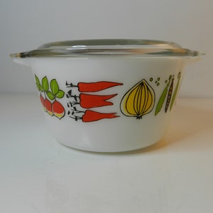 Op de afbeelding: Witte Pyrex-kom met een doorzichtig glazen deksel. De kom heeft een kleurrijk groentendesign, waaronder radijsjes, wortelen, een ui en erwten. Een vintage keukenartikel.