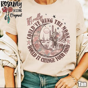May include: A tan t-shirt with a vintage-style graphic print of a cowboy riding a horse in front of a crescent moon and cacti. The text on the shirt reads "If I really could've hung the moon would it change your mind".