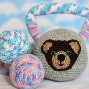 May include: A gray crocheted kettlebell with a brown bear face and green eyes. The kettlebell has a handle made of blue, pink, and white yarn. There are two smaller balls of yarn in the background, one blue, pink, and white, and the other pink, purple, and white.