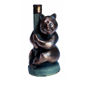 May include: A brown ceramic figurine of a bear holding a small, cylindrical object. The bear is sitting on a green base.