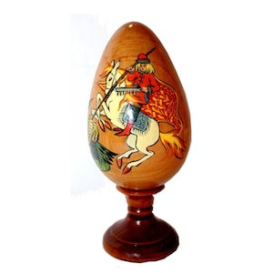 Puede incluir: Un adorno de madera con forma de huevo con una escena pintada de un caballero a caballo luchando contra un dragón. El adorno está sobre un soporte de madera.