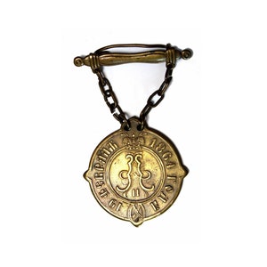 Puede incluir: Una medalla de metal dorado con una cadena y un cierre. La medalla tiene un diseño circular con el texto "1864 ГОДА" y "СЕГЕДЬ" alrededor del borde. El centro de la medalla presenta una corona y un águila bicéfala.