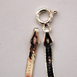 May include: Close-up of a silver chain necklace with a lobster clasp. The chain is a herringbone pattern and has a 925 stamp on the clasp.