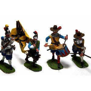 Puede incluir: Cuatro soldados de juguete en miniatura con uniformes militares del siglo XVII. Los soldados sostienen armas y una bandera con fondo amarillo y un águila negra. Los soldados están de pie sobre una base verde.