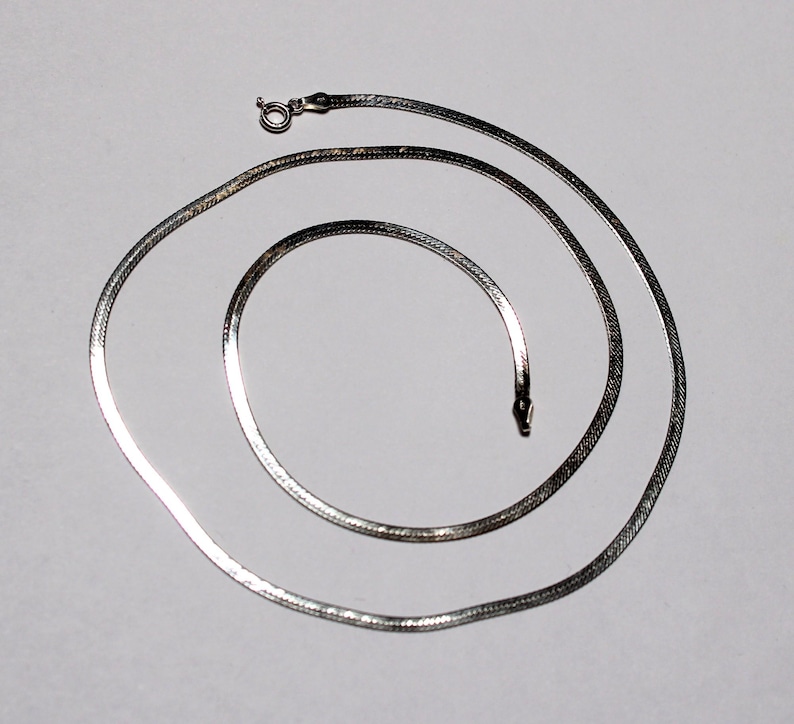 May include: A silver snake chain necklace. The chain is made of small, flat links that are woven together in a herringbone pattern.