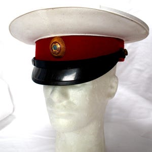 Op de afbeelding: Een witte militaire hoed met een rode band en een zwarte klep. De hoed heeft een gouden embleem aan de voorkant en wordt tentoongesteld op een mannequinhoofd. De rand van de hoed is wit en de klep is zwart.