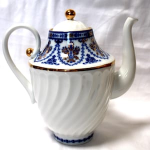 Puede incluir: Tetera de porcelana blanca con un diseño floral azul y dorado. La tetera tiene un patrón de remolino en el cuerpo y una perilla dorada en la tapa.