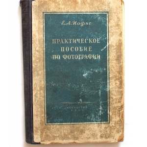 Puede incluir: Libro antiguo con una cubierta de color turquesa y bordes dorados. La portada presenta texto en ruso, incluido el título "Guía práctica de fotografía". El lomo del libro es azul oscuro.