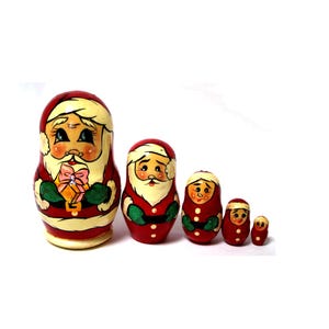 Könnte beinhalten: Satz von fünf roten und weißen russischen Nistpuppen, die den Weihnachtsmann darstellen. Die größte Puppe hält eine Geschenkbox mit einer rosa Schleife. Die Puppen sind mit Details wie weißem Bart, schwarzen Augen und grünem Hut bemalt.