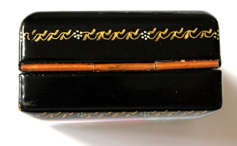 May include: A black lacquered wooden box with a gold painted design. The box has a hinged lid and a copper colored metal hinge.