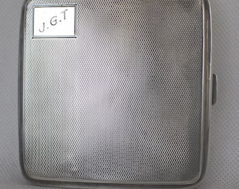 Op de afbeelding: Een zilverkleurige rechthoekige etui met een textuur. De etui heeft de initialen "J.G.T." in een klein rechthoekig kader. Afgeronde hoeken en een sluiting aan de zijkant.