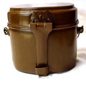 May include: A vintage olive-drab metal mess kit with a hinged lid and a folding handle. The kit has a cylindrical shape with a rounded top and bottom. The lid is a lighter shade of olive-drab. The kit is designed for outdoor use.