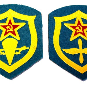 May include: Two embroidered patches with a yellow shield shape on a teal background. The shield contains a red star with a hammer and sickle inside, a yellow symbol, and a yellow winged symbol.