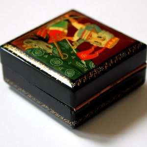 May include: A small black wooden box with a hinged lid. The box is decorated with a colorful folk art painting of a person in a red and white costume. The box has gold trim and a copper hinge.