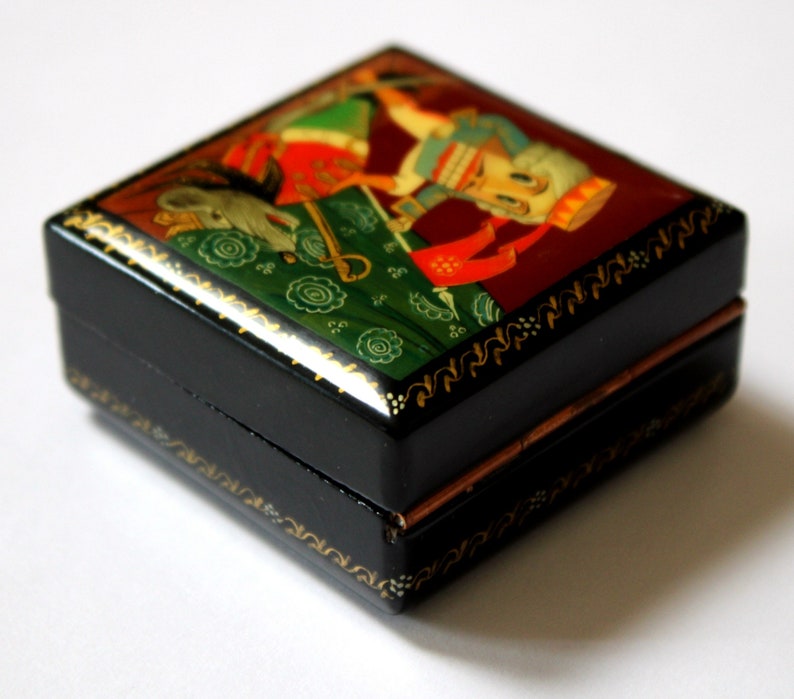 May include: A small black wooden box with a hinged lid. The box is decorated with a colorful folk art painting of a person in a red and white costume. The box has gold trim and a copper hinge.