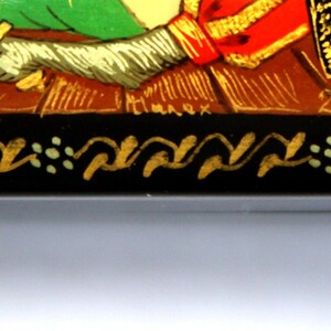 May include: A small wooden box with a black background and gold and green accents. The box is decorated with a painted scene of a knight on horseback. The box is signed "2002 -  Сарат."