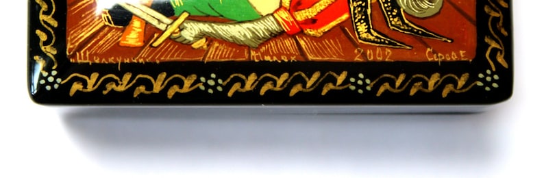 May include: A small wooden box with a black background and gold and green accents. The box is decorated with a painted scene of a knight on horseback. The box is signed "2002 -  Сарат."