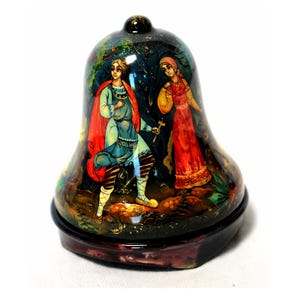 May include: A hand-painted wooden bell with a folk art design. The bell features a couple in traditional Russian clothing standing in a forest setting. The bell is decorated with intricate details and vibrant colors.