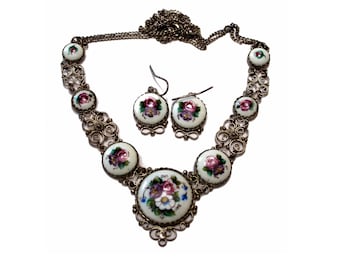 Peut inclure: Un collier en argent avec une chaîne délicate et des pendentifs ronds en porcelaine blanche ornés de motifs floraux roses, violets et verts. Des boucles d'oreilles assorties avec le même motif floral complètent l'ensemble.