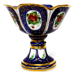 May include: A porcelain pedestal bowl featuring a deep blue and white color palette with hand-painted red rose motifs. The bowl is decorated with intricate gold filigree scrollwork and a scalloped rim edged in gold, set on a flared base.