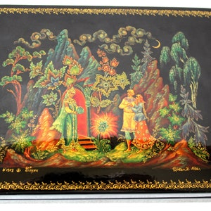 May include: A black lacquered wooden box with a painted scene of a couple standing in a forest with a magical portal. The scene is painted in vibrant colors with gold accents. The box is decorated with a gold border and the artist's signature is visible in the bottom right corner.