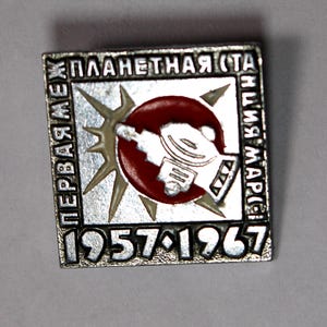 May include: A vintage square pin with a silver border and Russian text. The pin features a red circle with a white illustration of a spacecraft, surrounded by a starburst design. The years 1957-1967 are displayed at the bottom.