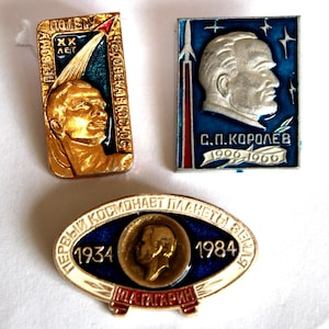 Puede incluir: Tres insignias vintage con temática espacial. Una insignia dorada presenta un retrato y texto en ruso. Una insignia azul tiene un cohete y un retrato plateado con el texto "C.P. Korolev 1906-1966". La tercera insignia tiene un retrato y el texto "1934-1984".