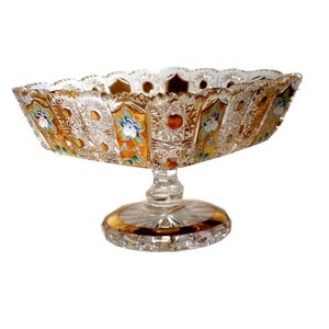 May include: A square-shaped crystal pedestal bowl featuring intricate cut glass patterns, amber-colored glass panels, and hand-painted floral motifs in blue, green, and white. The bowl stands on a clear glass base with a gold-toned rim accent.