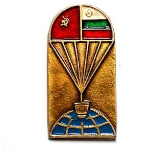 Peut inclure: Épingle vintage avec un marteau et une faucille rouges, un drapeau avec des rayures vertes et blanches et un motif de parachute. L'épingle a un fond doré et un globe bleu en bas.