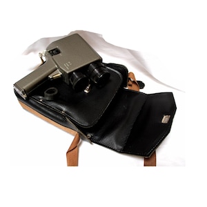May include: A vintage silver-grey film camera with two black lenses sits in an open black leather case with a tan border and brown straps. The camera is angled towards the viewer, showcasing its design.