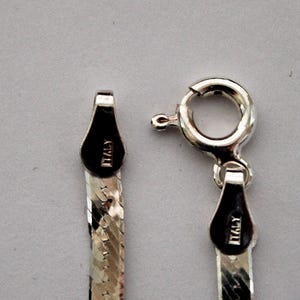 May include: Close-up of a silver chain necklace with a lobster clasp. The chain is made of small, flat links and has a diamond-cut finish. The clasp has a small tag that reads "ITALY".