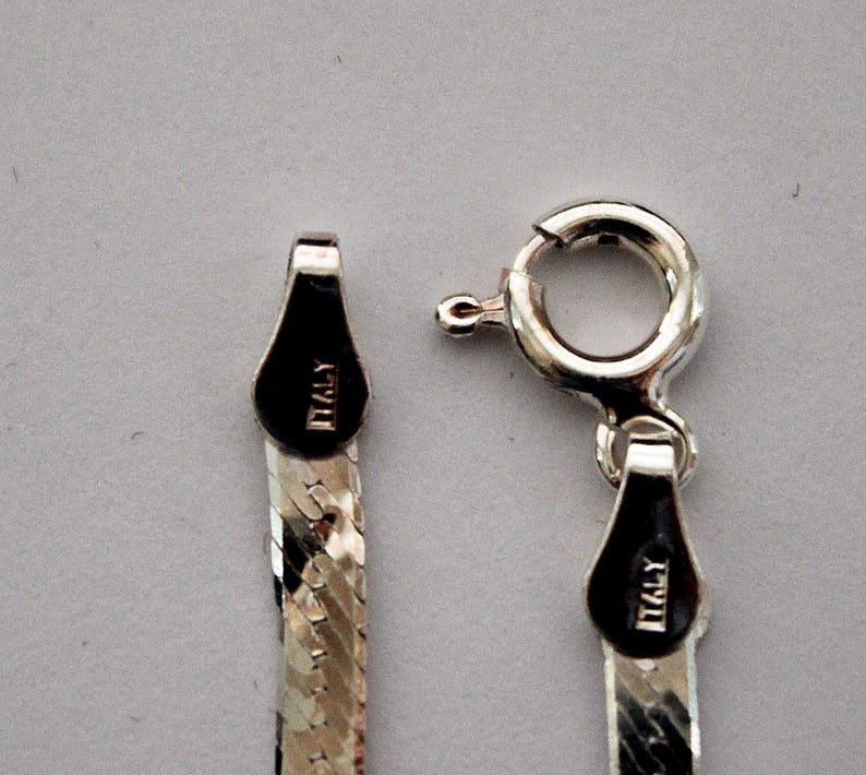 May include: Close-up of a silver chain necklace with a lobster clasp. The chain is made of small, flat links and has a diamond-cut finish. The clasp has a small tag that reads "ITALY".