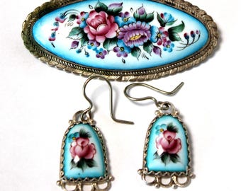 Peut inclure: Un ensemble de boucles d'oreilles et d'une broche en argent avec émail bleu et motifs floraux peints à la main. Les boucles d'oreilles sont des boucles d'oreilles pendantes avec un pendentif en forme de larme. La broche est de forme ovale.