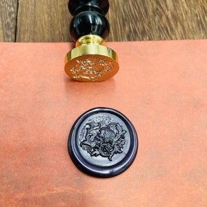May include: A black and gold wax seal stamp with a detailed floral design. The stamp is used to create a wax seal with a dark purple wax impression.