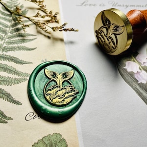 Op de afbeelding: Een groene waszegel met een gouden walvisstaartontwerp. De zegel bevindt zich op een witte achtergrond met een bloemenpatroon.