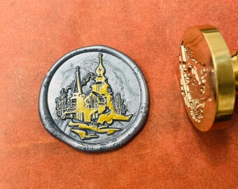 May include: A silver wax seal with a gold design of a church building with a steeple and trees. The wax seal is on a red surface.