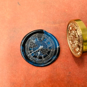 Può includere: Un sigillo di cera blu con un designo dettagliato di quadrante dell'orologio. Il sigillo è fatto di cera e ha un bordo dorato.