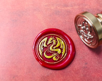 Könnte beinhalten: Ein rotes Wachssiegel mit einem goldenen Drachenmotiv. Das Siegel ist rund und hat einen erhöhten Rand. Der Drache ist nach rechts gerichtet und hat seine Flügel ausgebreitet.