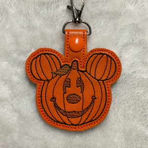 Puede incluir: Llavero naranja con forma de calabaza con detalles cosidos en negro y un cierre plateado. La calabaza tiene orejas de Mickey Mouse y una cara sonriente de linterna. Una pequeña lengüeta naranja con un botón a presión conecta la calabaza al cierre.