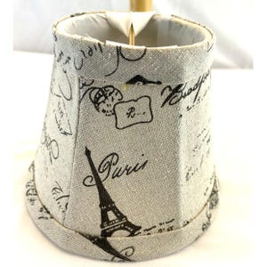 May include: A small, beige lampshade with a black Eiffel Tower illustration and the word "Paris" printed on the front. The shade is decorated with other black script and design elements.