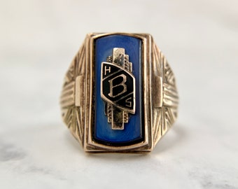 Könnte beinhalten: Ein goldener Klassenring mit einem blauen Stein und einem schwarzen und goldenen Emblem mit der Aufschrift "H.S. B".