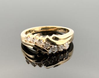 Puede incluir: Un anillo de oro con un diseño de onda, engastado con múltiples diamantes pequeños. El anillo se refleja en una superficie oscura y brillante, creando un efecto de espejo. El anillo está marcado con "14K".